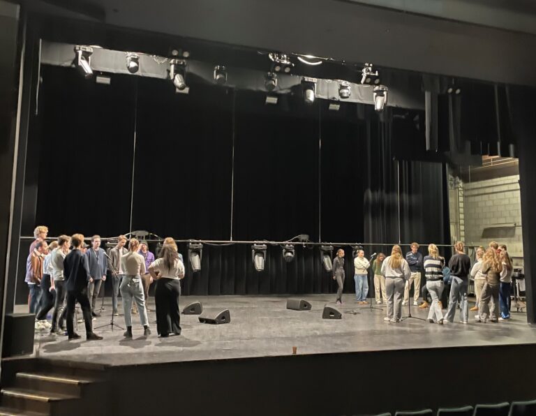 Een groep mensen staat op een toneel met zwarte gordijnen en theaterverlichting.