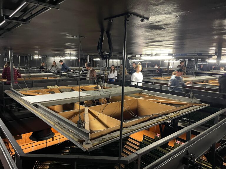 Een blik op een industrieel plafond met een hangend platform en mensen die zich eromheen bewegen.