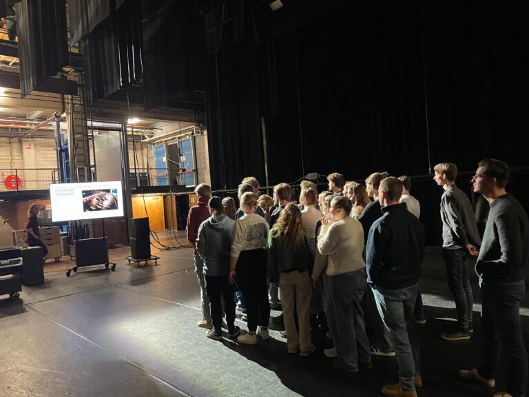 Een groep mensen staat op het podium met een scherm op de achtergrond, zichtbaar in een theater.