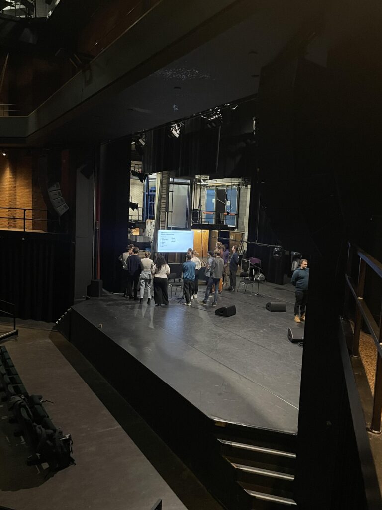 Een groep mensen staat op het podium van een theater tijdens een repetitie of evenement.