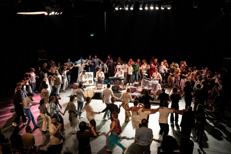 Een grote groep mensen danst in een kring de Dabke, hand in hand, met een live band in het midden van de kring op een verlicht podium.
