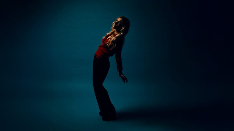 Agnes Obel staat voor een blauwe fotomuur. Ze buigt zich scheef achterover. Ze heeft een rode top en een zwarte broek aan en kijkt in een schijnwerper