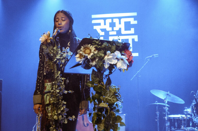 Een zangeres staat op het podium met een microfoon, omringd door een bloemenversiering, terwijl ze zingt voor een publiek.