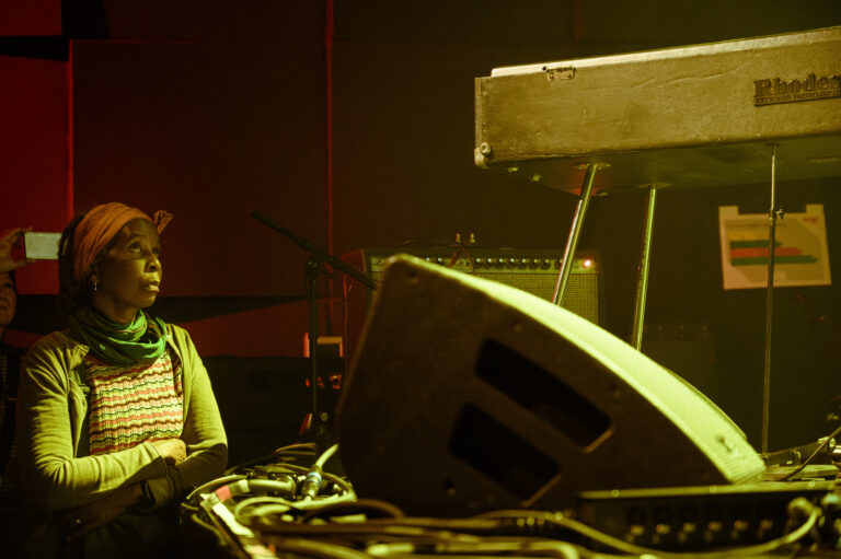Een vrouw kijkt aandachtig naar een keyboard en geluidssysteem in een sfeervolle muzieksetting.