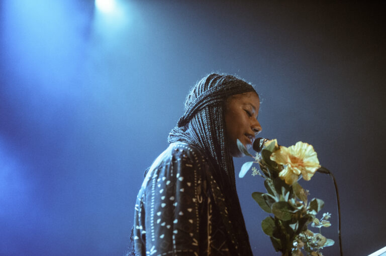 Een artiest met vlechten zingt in een microfoon, terwijl ze een gele roos vasthoudt tegen een blauw verlichte achtergrond.