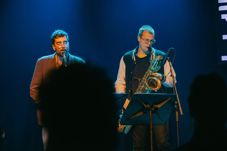 Twee muzikanten staan op een podium, één zingt terwijl de ander een saxofoon bespeelt, met een blauwe achtergrondverlichting.