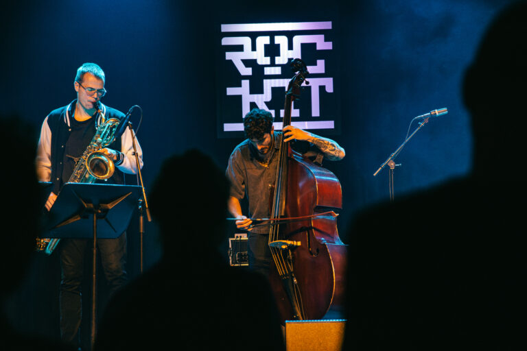 Twee muzikanten voeren live jazzmuziek uit op het podium, met een saxofonist en een contrabassist.