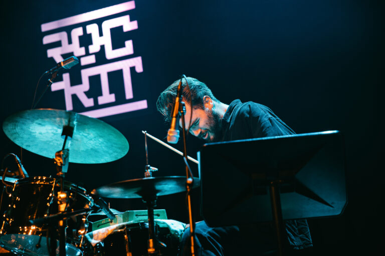 Een drummer speelt op een podium, met een verlichte achtergrond waarin de tekst