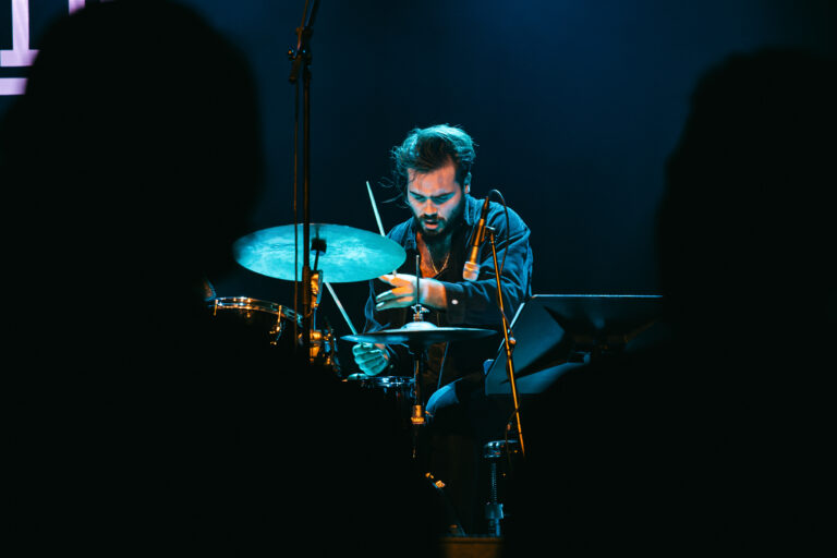 Een drummer speelt geconcentreerd op een podium, met het publiek op de achtergrond.