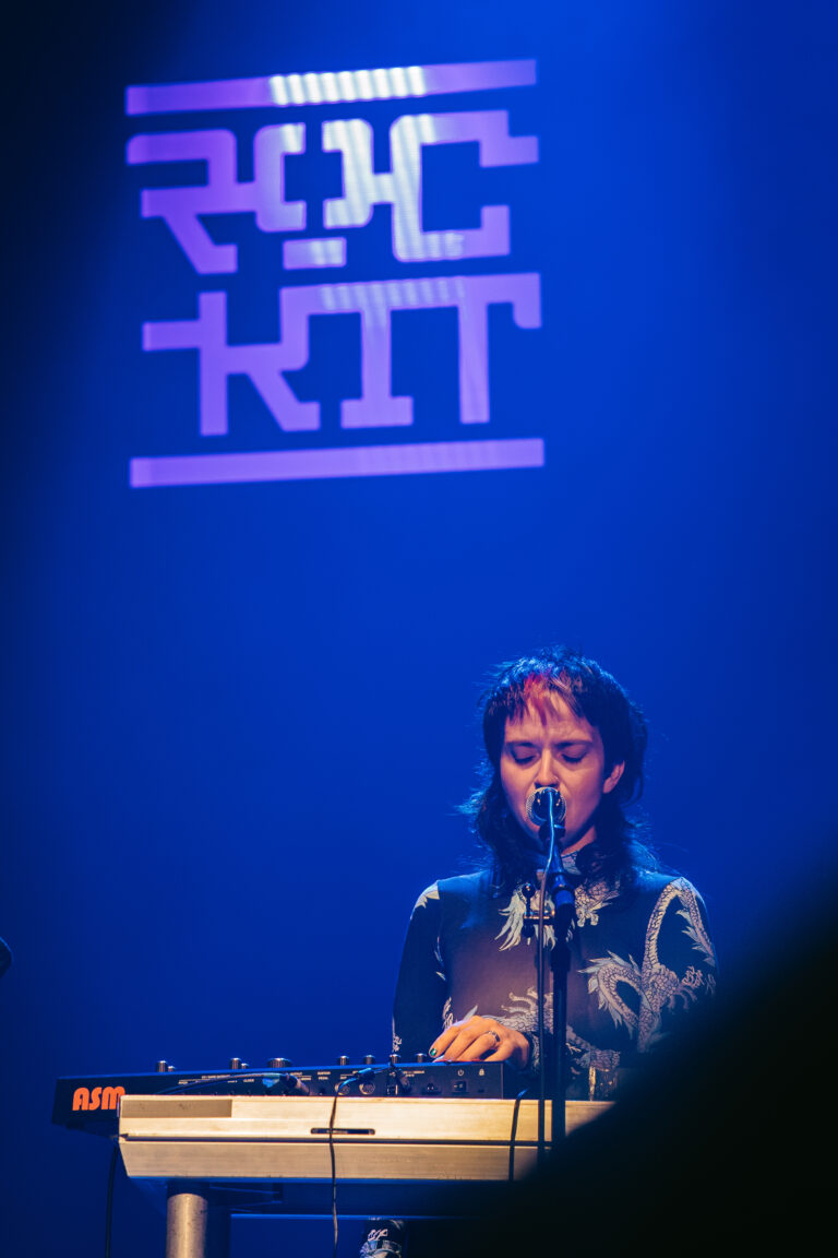 Een muzikante staat achter een synthesizer en zingt, met een verlichte achtergrond met de tekst