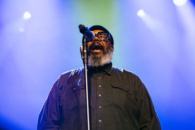 Een man met een baard staat naast een microfoon en zingt op een podium onder sfeervolle verlichting.
