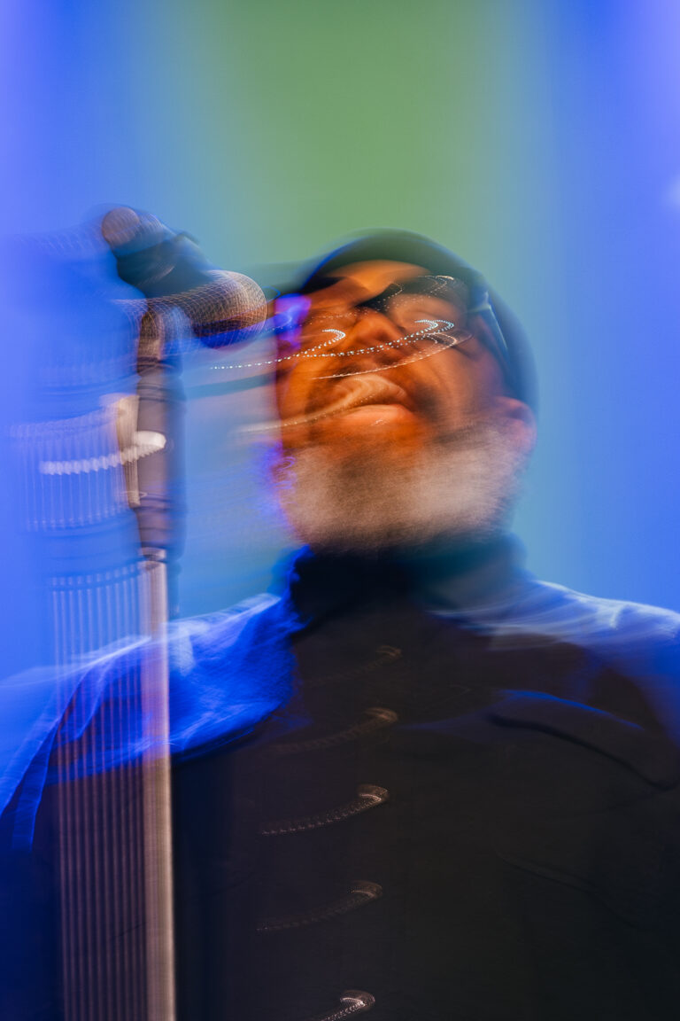 Een zanger op het podium met een microfoon, omlijst door gekleurde lichten en een onscherpe beweging.