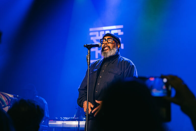 Een man met een grijze baard zingt in een sfeervol verlichte club, terwijl het publiek hem vanuit de schaduw observeert.