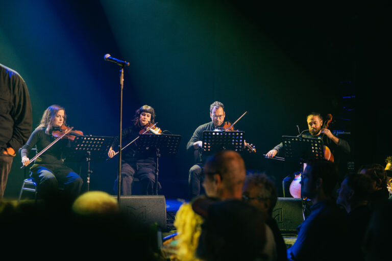 Een strijkkwartet speelt op een podium terwijl het publiek in de schaduw toekijkt.