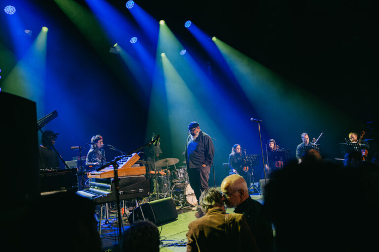 Een live muziekevenement met een band op het podium, verlicht door blauwe en groene lichten.