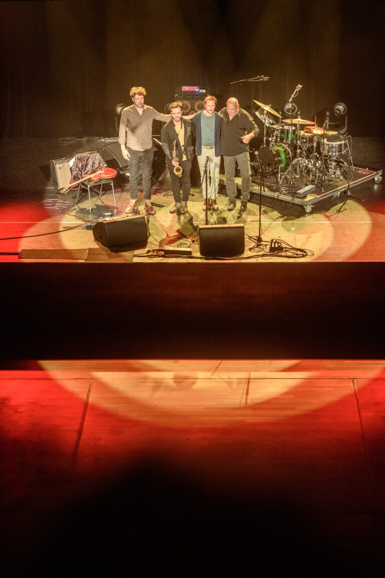 Een muzikale band poseert op een podium met instrumenten in de achtergrond en gekleurd licht.