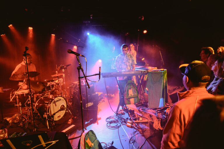 Een band speelt live op het podium terwijl het publiek enthousiast toekijkt, omgeven door kleurrijke lichten.
