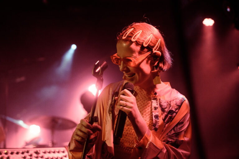 Een performer met een kleurrijke outfit staat op een podium met een microfoon en een vrolijke uitstraling, omgeven door sfeervolle lichtjes.