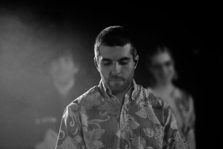 Een persoon in een bloemenmotief overhemd staat voor een donkere achtergrond met vage silhouetten van andere mensen.