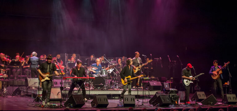 Een groot muziekensemble speelt op een podium met kleurrijke lichteffecten.