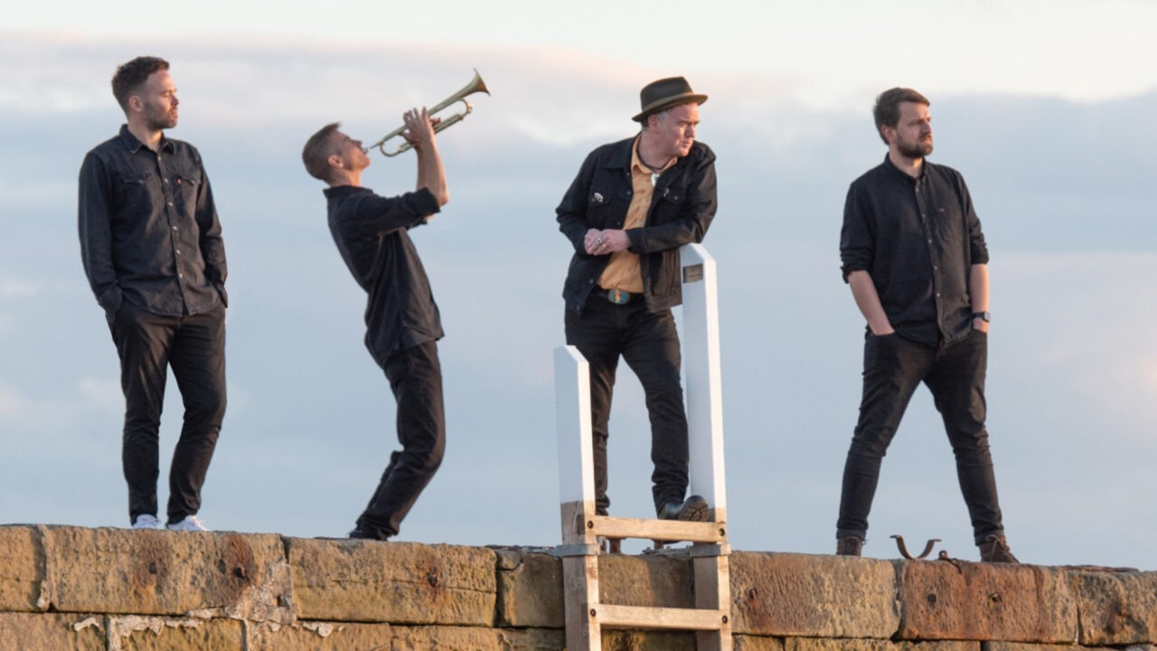 De bandleden van Dean Owens & the Sinners poseren op een stenen muur tegen een blauwe lucht, met een trompettist die zijn instrument omhoog houdt en de anderen ontspannen in zwart gekleed.