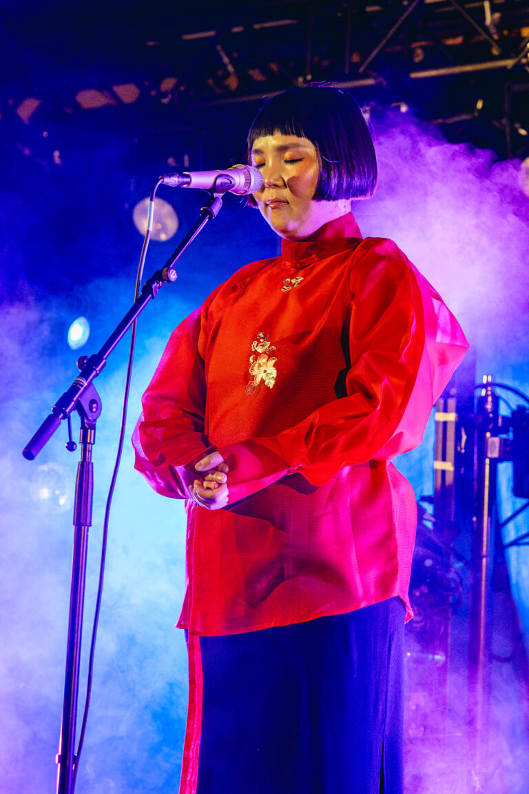 Een zangeres in traditionele kleding staat voor een microfoon, omringd door kleurrijke lichtsferen en rook op het podium.