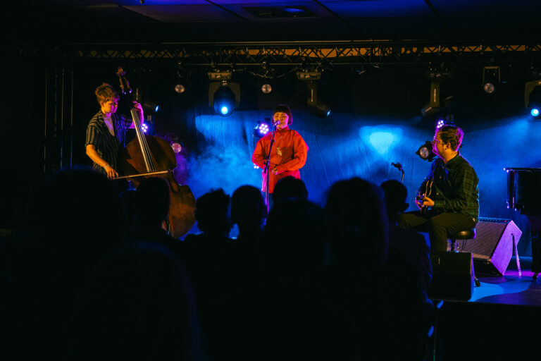 Een muzikale liveoptreden met een zanger in het rood en muzikanten op een donkere podium met blauwe verlichting.
