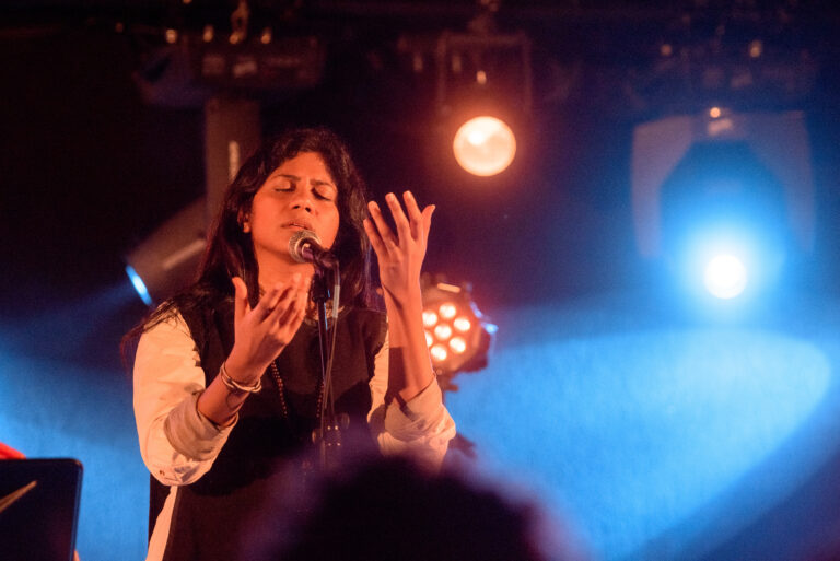 Een zangeres staat op een podium en maakt een emotionele gesticuleerende beweging terwijl ze zingt, omringd door kleurrijke lichten.