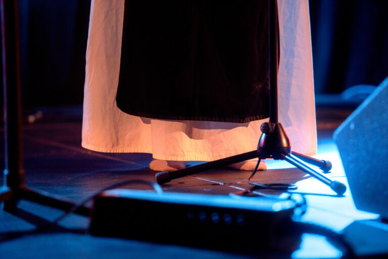 Een close-up van een podium met een rok, een microfoonstandaard en een effectenpedaal verlicht door verschillende kleuren lampen.