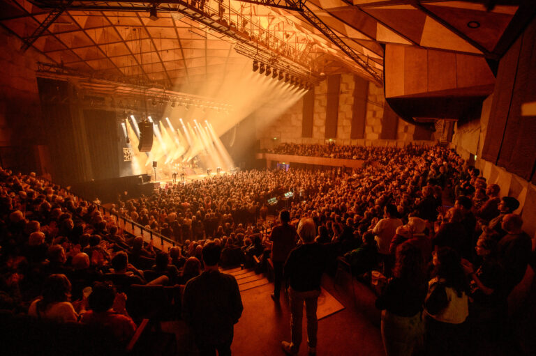 Een druk concert met een volle zaal, waar het podium verlicht is door grote lichtstralen.