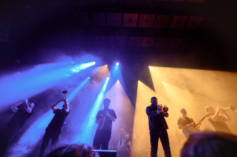 Een groep muzikanten staat op een podium onder kleurrijke lichten tijdens een live optreden.
