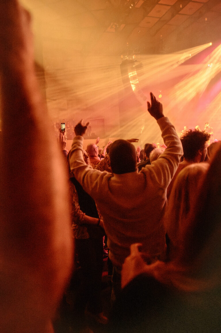Een menigte mensen geniet van een optreden met opgestoken handen in een sfeervolle, verlichte ruimte.