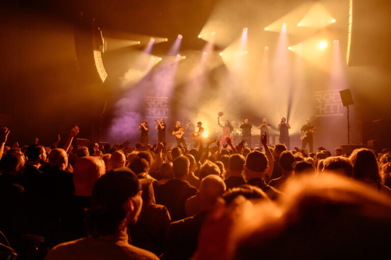 Een druk concert met een groep performers op het podium, omringd door een enthousiast publiek onder fel licht.