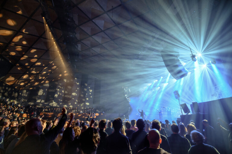 Een drukke concertzaal met een uitzinnig publiek en kleurrijke lichtstralen die vanaf het podium stralen.