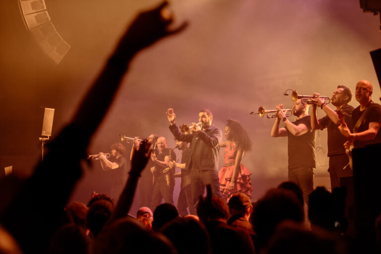 Een muzikale uitvoering met een zanger en blazers, omringd door een enthousiast publiek met opgestoken handen in een sfeervolle lichtsetting.