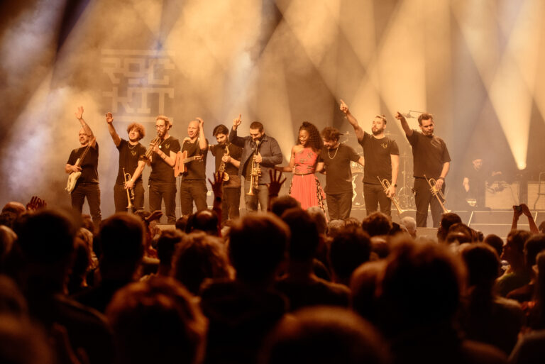 Een groep muzikanten staat geïllumineerd op het podium, terwijl het publiek voor hen juicht.