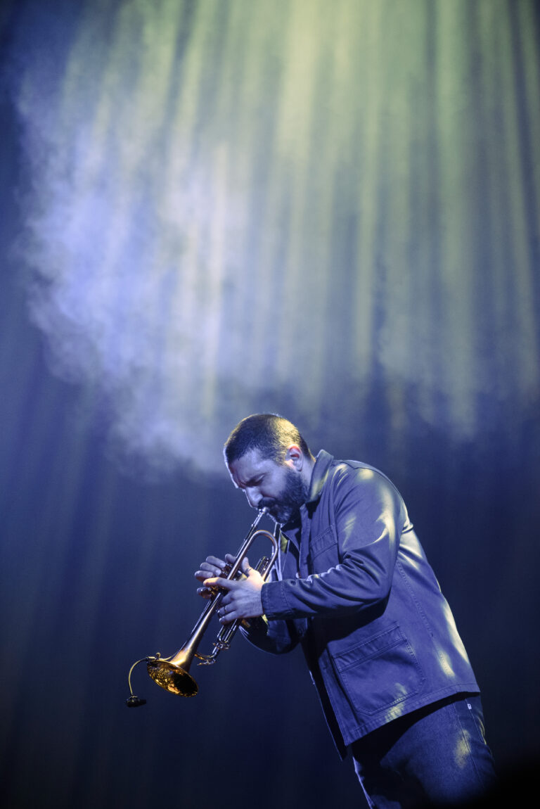 Een muzikant speelt trompet onder een spotlight met rook en lichtstralen op de achtergrond.