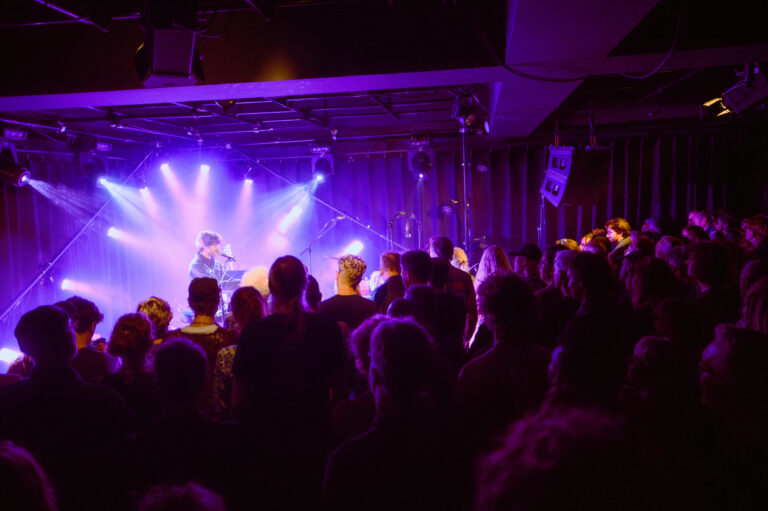 Een menigte geniet van een live optreden met paarse verlichting in een zaal.