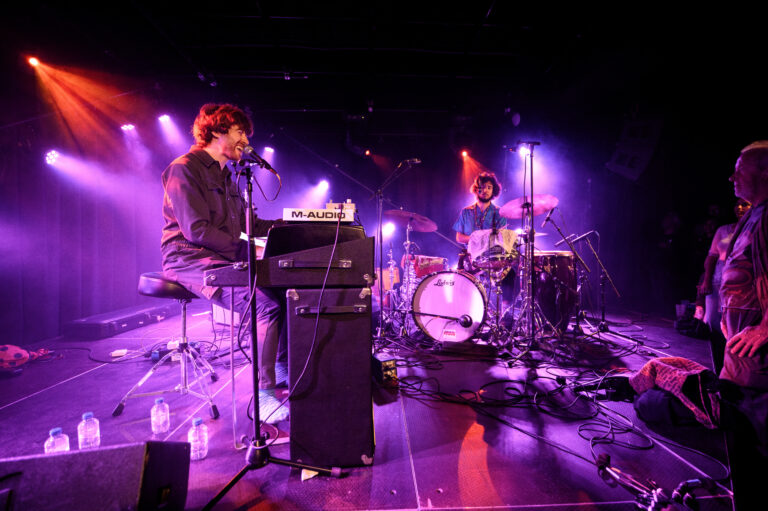 Een muzikant speelt keyboard en zingt op een podium, verlicht door paarse en oranje lichten, terwijl een drummer naast hem speelt.