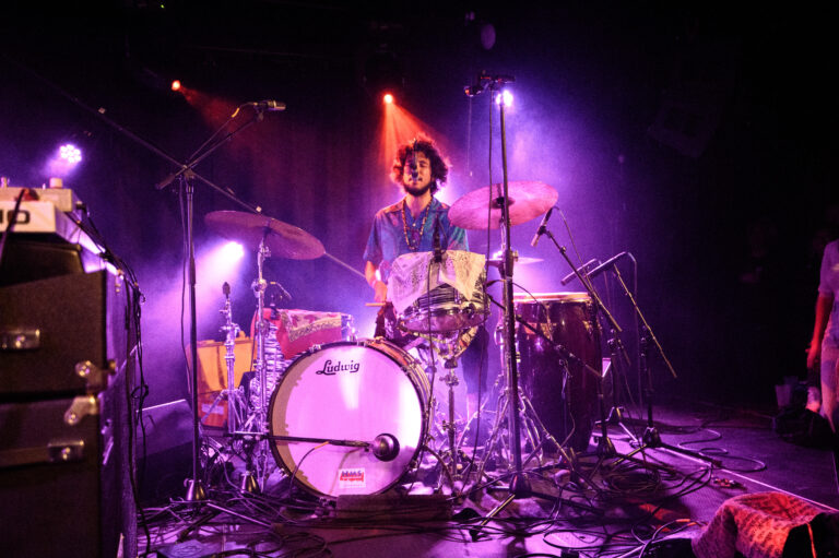 Een drummer speelt op een podium omringd door kleurrijke lichten.