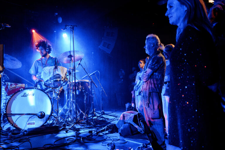 Een drummer speelt op een podium terwijl een klein publiek onder een blauwe lichtshow toekijkt.