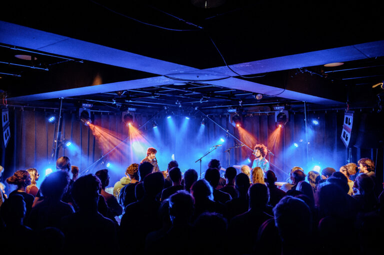 Een live muziekoptreden met een energiek publiek in een donkerverlichte zaal met kleurrijke verlichting.