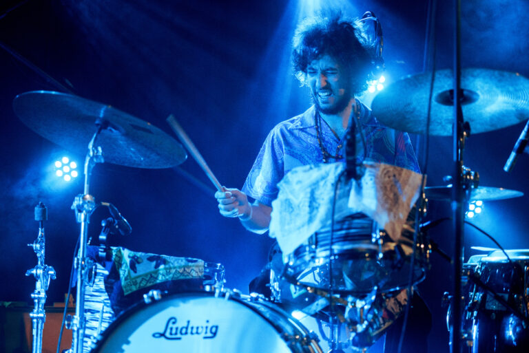 Een drummer speelt energiek op een set drums, verlicht door sfeervolle blauwe lichten.