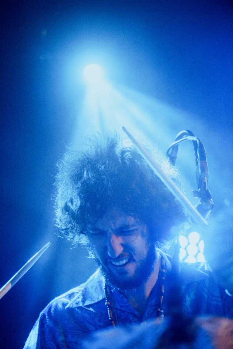 Een drummer speelt intensief in een sfeervolle, verlichte omgeving met blauwe lichten.