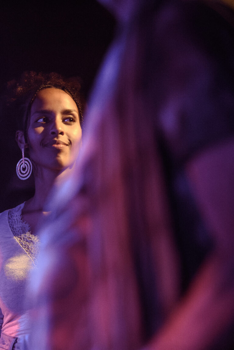 Een vrouw met een glimlach en grote oorbellen staat in een sfeervolle, kleurrijke achtergrond.