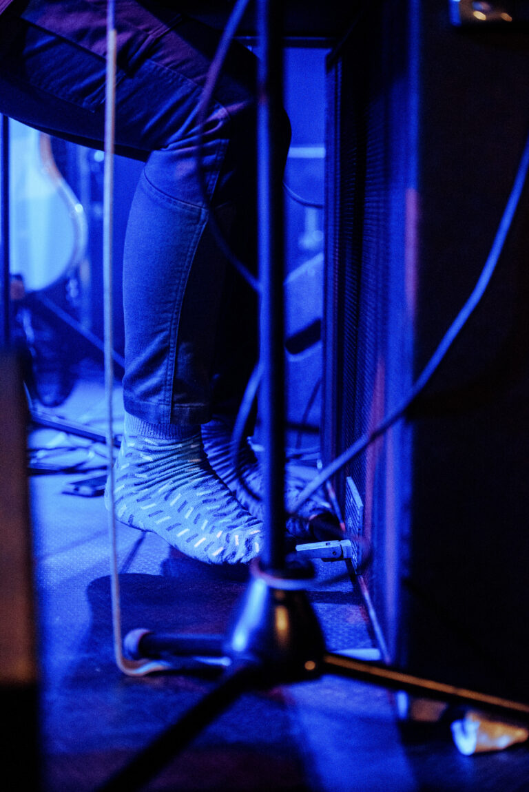 Een close-up van een persoon die speelt op een instrument, met focus op hun voeten en de apparatuur eromheen, verlicht in blauwe tonen.