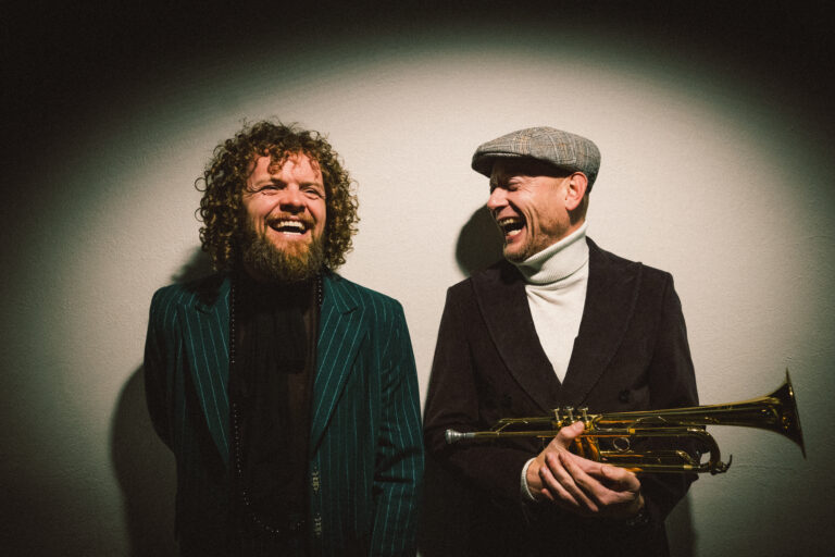 Voor een spotlight staan Marcel Veenendaal en Jan van Duikeren te lachen. Jan heeft een instrument vast en kijkt naar Marcel.