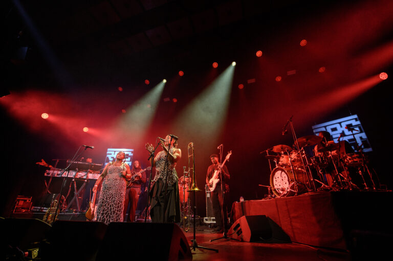Een band staat op het podium tijdens een optreden, met kleurrijke lichten en muziekinstrumenten zichtbaar.