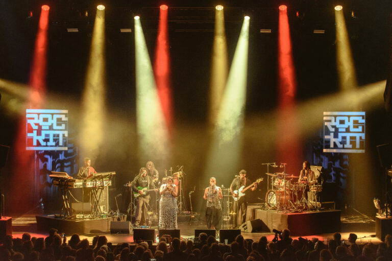 Een band treedt live op met diverse muzikanten en lichteffecten op een podium.