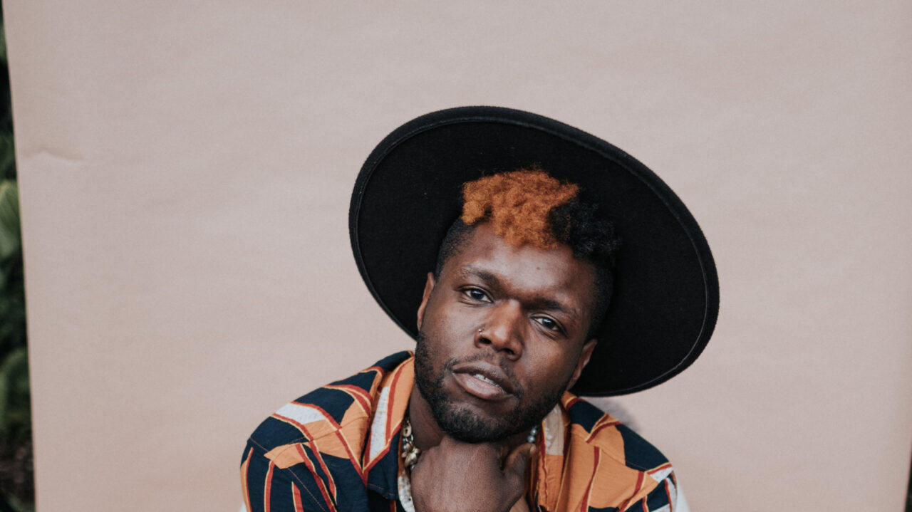 Portret van Les Greene met een grote zwarte hoed, oranje-zwart gekleurd haar, en een opvallend gestreept shirt, die ontspannen in de camera kijkt.